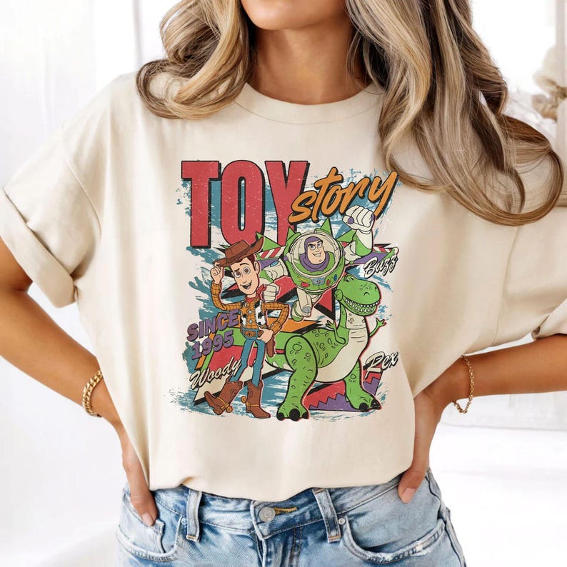 May include: A cream-colored short-sleeve t-shirt featuring a graphic print of Woody, Buzz Lightyear, and Rex from Toy Story. The design includes the text "TOY story", "SINCE 1995", "Woody", "Buzz", and "Rex" in a colorful, distressed artistic style.