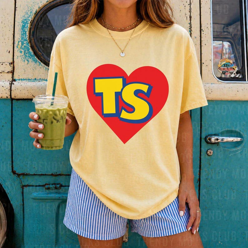 K&ouml;nnte beinhalten: Ein blassgelbes Kurzarm-T-Shirt mit einem gro&szlig;en roten Herz-Motiv auf der Brust. Im Inneren des Herzens sind die Buchstaben TS in einer kr&auml;ftigen gelben Schrift mit blauem Umriss abgebildet. Das T-Shirt wird mit blau-wei&szlig; gestreiften Shorts kombiniert.