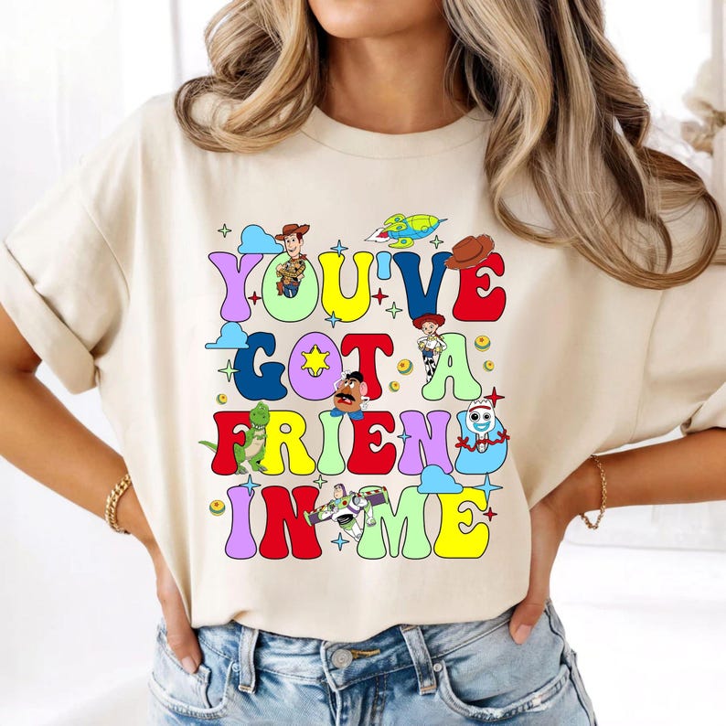 K&ouml;nnte beinhalten: Ein cremefarbenes T-Shirt mit einem bunten Grafikdruck, der den Text "YOU'VE GOT A FRIEND IN ME" in gro&szlig;en, blasenartigen, mehrfarbigen Buchstaben zeigt. Das Design enth&auml;lt verschiedene Zeichentrickfiguren aus Toy Story, darunter Woody, Jessie, Rex, Mr. Kartoffelkopf, Forky und Buzz Lightyear, sowie kleine Stern- und Wolkenakzente.