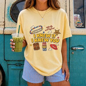 May include: A pale yellow short-sleeve t-shirt featuring a graphic print with the text "I KNEW IT I KNEW YOU", "infinity and beyond", and "YOU'VE GOT A FRIEND IN ME". The design includes illustrations of cowboy boots, a red hat, and alphabet blocks.