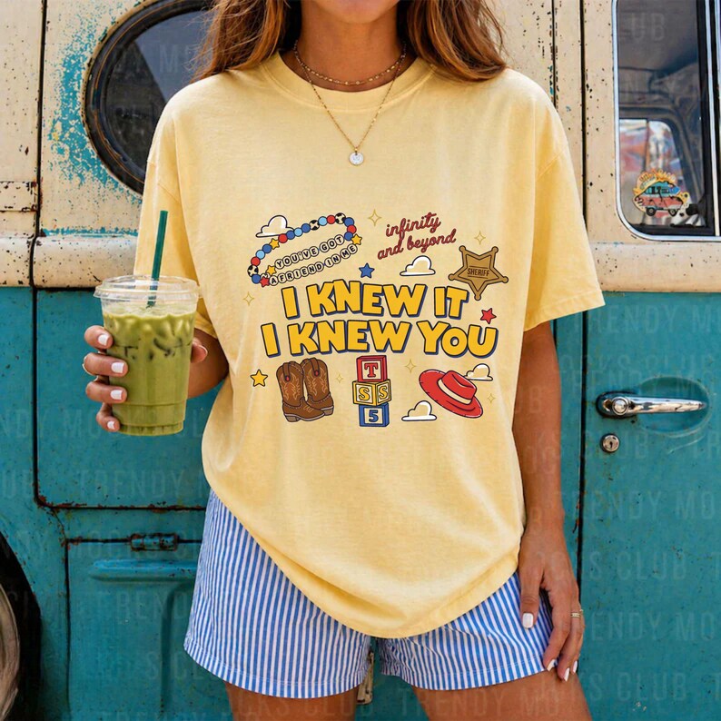 May include: A pale yellow short-sleeve t-shirt featuring a graphic print with the text "I KNEW IT I KNEW YOU", "infinity and beyond", and "YOU'VE GOT A FRIEND IN ME". The design includes illustrations of cowboy boots, a red hat, and alphabet blocks.