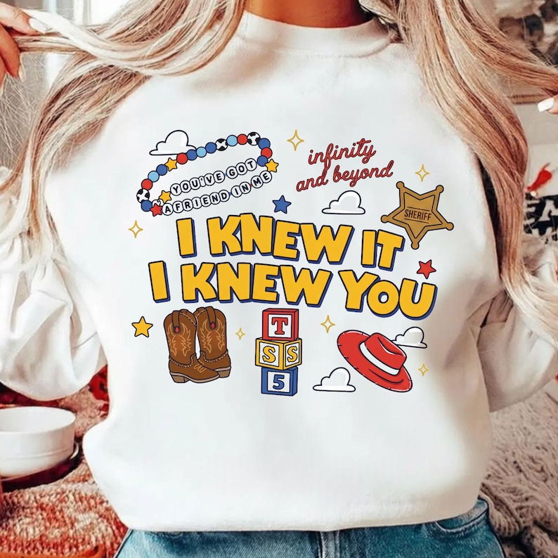 K&ouml;nnte beinhalten: Ein wei&szlig;es Sweatshirt mit Rundhalsausschnitt und einem Grafikdruck mit dem gelben Text I KNEW IT I KNEW YOU, einem Perlenarmband mit dem Text YOU'VE GOT A FRIEND IN ME, einem Sheriffstern, Cowboystiefeln, Buchstabenw&uuml;rfeln und einem roten Hut.