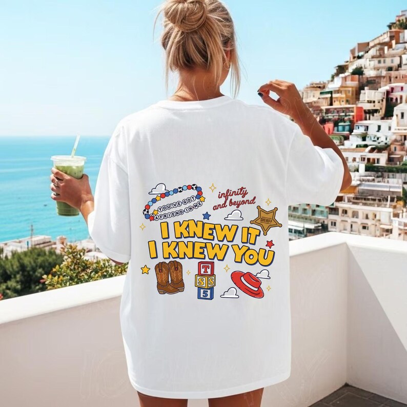 May include: A white t-shirt featuring a colorful graphic on the back with the text "I KNEW IT I KNEW YOU", "infinity and beyond", and "YOU'VE GOT A FRIEND IN ME". The design includes illustrations of cowboy boots, a sheriff badge, and blocks.