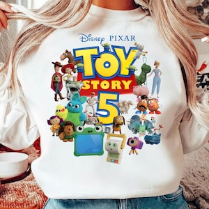 K&ouml;nnte beinhalten: Ein wei&szlig;es Langarm-Sweatshirt mit einem farbenfrohen Grafikdruck des Disney Pixar Toy Story 5-Logos, umgeben von verschiedenen animierten Charakteren wie Woody, Buzz Lightyear, Jessie, Rex, Slinky Dog, Mr. und Mrs. Potato Head und anderen.