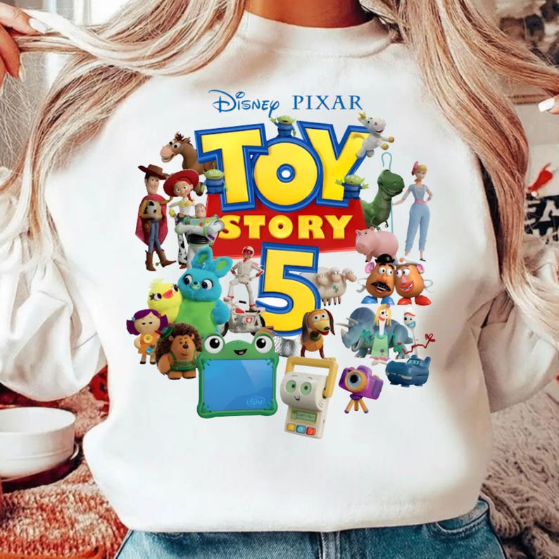K&ouml;nnte beinhalten: Ein wei&szlig;es Langarm-Sweatshirt mit einem farbenfrohen Grafikdruck des Disney Pixar Toy Story 5-Logos, umgeben von verschiedenen animierten Charakteren wie Woody, Buzz Lightyear, Jessie, Rex, Slinky Dog, Mr. und Mrs. Potato Head und anderen.