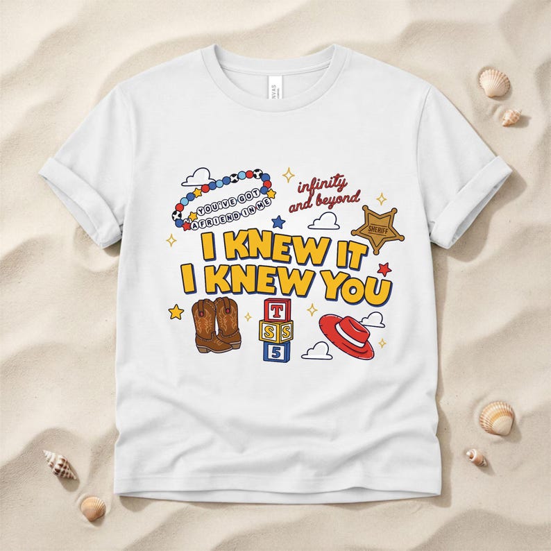 May include: A white short-sleeve t-shirt featuring a colorful graphic print with the text "I KNEW IT I KNEW YOU", "infinity and beyond", and "YOU'VE GOT A FRIEND IN ME". The design includes illustrations of cowboy boots, a sheriff badge, and blocks.