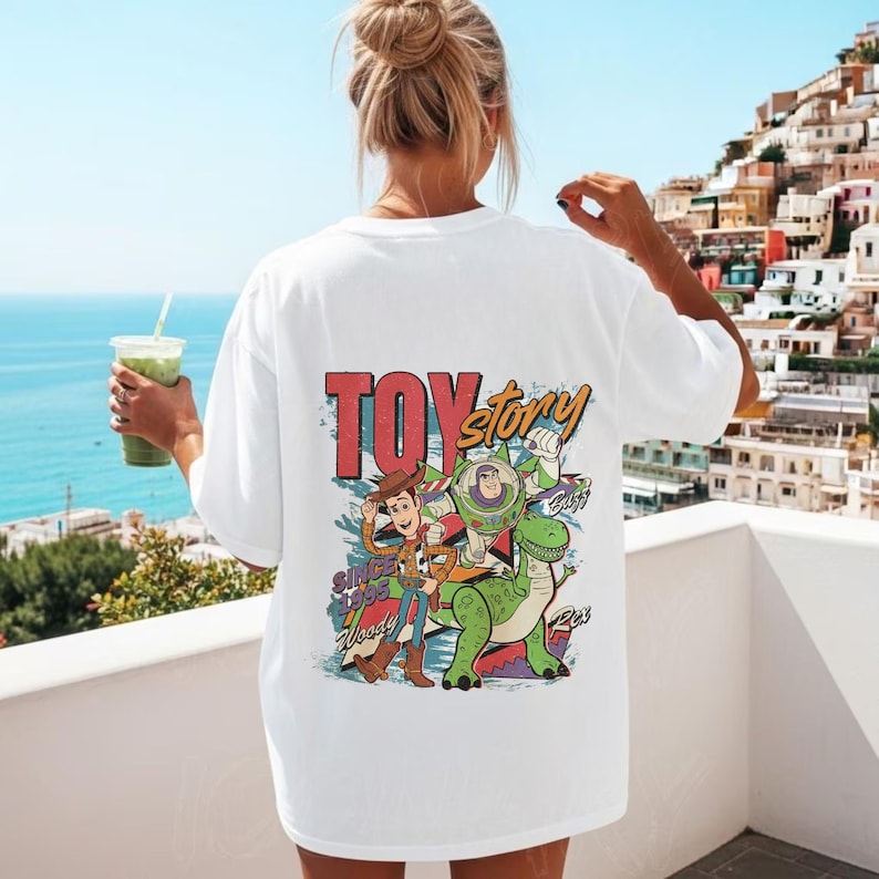 May include: A white short-sleeve t-shirt featuring a colorful graphic on the back. The print displays the text Toy Story, Since 1995, Woody, Buzz, and Rex. The design includes illustrations of the characters Woody, Buzz Lightyear, and Rex.