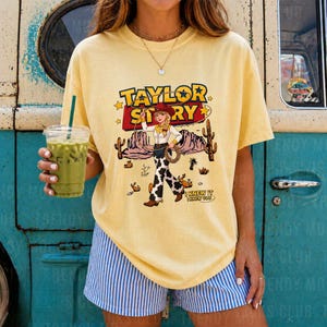 K&ouml;nnte beinhalten: Ein blassgelbes T-Shirt mit kurzen &Auml;rmeln und einem Grafikdruck einer Cowgirl mit Lasso. Der Text lautet TAYLOR STORY und I KNEW IT I KNEW YOU. Das Design enth&auml;lt W&uuml;stenelemente wie Kakteen und Sterne in einer westlich inspirierten Illustration.
