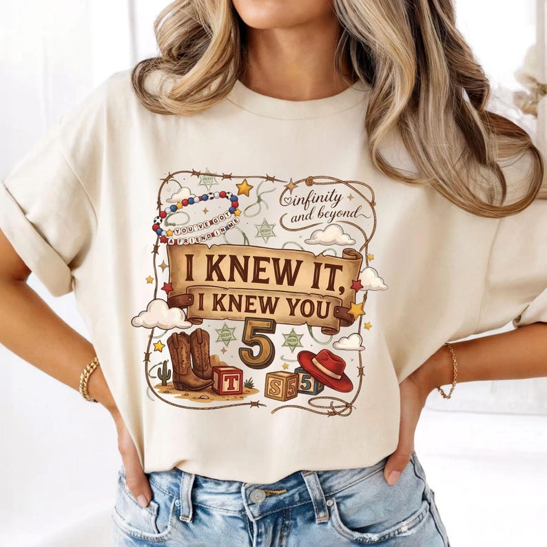 May include: A cream-colored t-shirt featuring a graphic print with a brown scroll banner that reads "I KNEW IT, I KNEW YOU" above the number "5". The design includes a cowboy boot, a red hat, alphabet blocks, a lasso, and "Infinity and beyond".