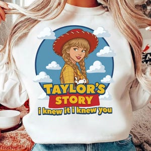 Op de afbeelding: Een wit sweatshirt met een afbeelding van een stripfiguur met blond gevlochten haar, een rode cowboyhoed en een geel geruit overhemd. Het ontwerp bevat een blauwe lucht met witte wolken en de tekst Taylor's Story en i knew it i knew you.
