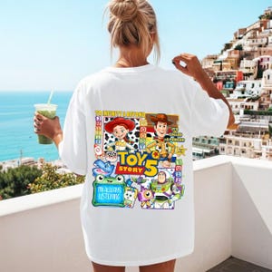 May include: A white t-shirt featuring a colorful graphic on the back with Toy Story characters including Jessie, Woody, and Buzz Lightyear. The design includes the text To Infinity & Beyond, Toy Story 5, and I'm Always Listening, along with various icons.