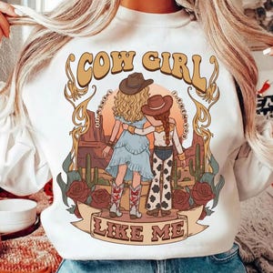 K&ouml;nnte beinhalten: Ein wei&szlig;es Sweatshirt mit einem Grafikdruck von zwei Figuren mit Cowboyh&uuml;ten, die in einer W&uuml;stenlandschaft mit Kakteen und Rosen stehen. Der Text COW GIRL ist oben und LIKE ME unten in einer beigen Retro-Schriftart aufgedruckt.