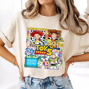 May include: A beige short-sleeve t-shirt featuring a colorful graphic print of Toy Story characters including Jessie, Woody, and Buzz Lightyear. The design includes the text TO INFINITY & BEYOND, TOY STORY 5, IT'S CALLED A PONCHO, and I'M ALWAYS LISTENING.