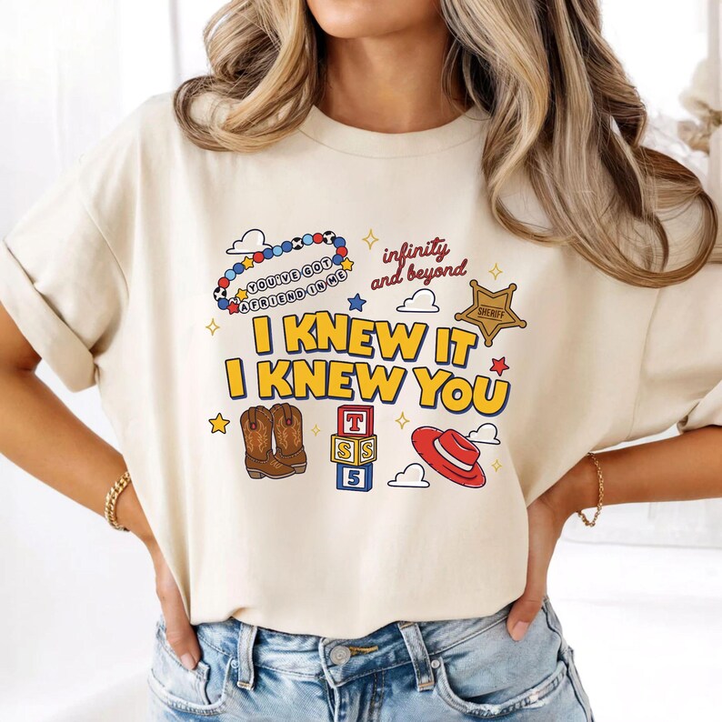 May include: A cream-colored t-shirt featuring a graphic print with the text "I KNEW IT I KNEW YOU", "YOU'VE GOT A FRIEND IN ME", and "infinity and beyond". The design includes illustrations of a beaded necklace, a sheriff badge, cowboy boots, blocks, and a hat.