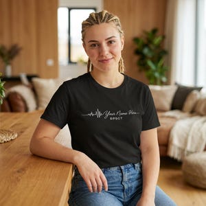 Op de afbeelding: Zwart t-shirt met een wit hartslagontwerp en de tekst "Your Name Here" en "RPSGT". Het shirt is gemaakt van een zacht materiaal en heeft een klassieke ronde hals.