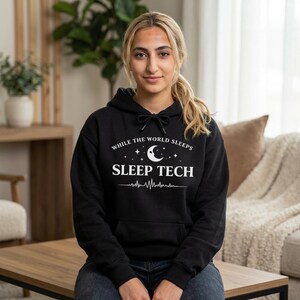 May include: Black hoodie featuring the white text "WHILE THE WORLD SLEEPS SLEEP TECH". The design includes a crescent moon, stars, and a heartbeat line. The hoodie has a drawstring hood and a front pocket.