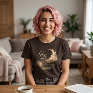 Könnte beinhalten: Ein braunes T-Shirt mit dem Text "Sleep Tech" in goldener Schrift, einem Halbmond und einem floralen Design. Das Shirt hat auch den Text "Monitoring Dreams Professionally". Die Person auf dem Bild lächelt.
