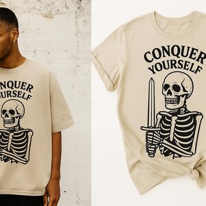 Pode incluir: Camiseta bege com a frase "CONQUER YOURSELF" acima de uma ilustração em preto e branco de um esqueleto segurando uma espada. A camiseta é de cor neutra e tem mangas curtas.