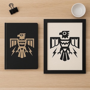 May include: A black notebook and framed print featuring a gold and black stylized eagle design with lightning bolts. The notebook is closed and the print is in a black frame. A small white bowl and a black binder clip are also visible.