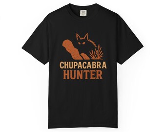 May include: Black t-shirt with a graphic featuring a stylized Chupacabra illustration in brown and orange tones. The text "CHUPACABRA HUNTER" is printed below the image, making it a unique apparel item.