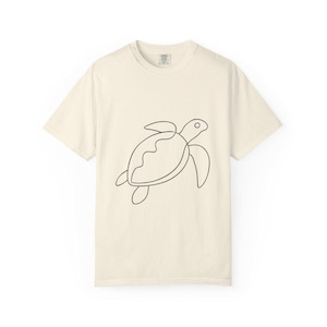 May include: A cream-coloured t-shirt with a minimalist black outline of a sea turtle. The design shows the turtle's shell, flippers, and head. The shirt is made of a soft material and has a classic crew neck.