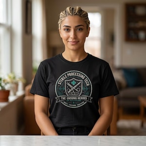 Op de afbeelding: Zwart T-shirt met een cirkelvormige afbeelding met de tekst "STERILE PROCESSING TECH" met een kruis en schaar ontwerp. De tekst "THE UNSUNG HEROES OF HEALTHCARE" staat eronder. Het T-shirt wordt gedragen door een persoon met gevlochten haar.