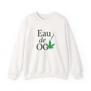 May include: White crew neck sweatshirt featuring the text "Eau de OG" in black, and a green cannabis leaf graphic. A casual style sweatshirt suitable for everyday wear.