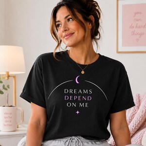 Peut inclure: Un t-shirt noir à manches courtes arborant un motif graphique composé d'un croissant de lune, d'une ligne courbe et du texte "DREAMS DEPEND ON ME" en lettres blanches et violet clair, avec une petite étoile à quatre branches en dessous.