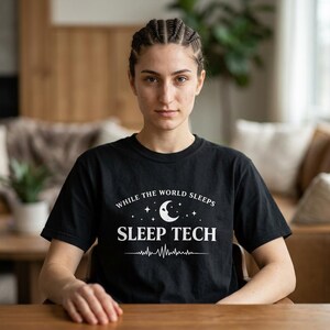 Peut inclure: T-shirt noir avec l'inscription blanche "WHILE THE WORLD SLEEPS SLEEP TECH". Le motif comprend un croissant de lune, des étoiles et une ligne de battement de cœur. Le t-shirt est à manches courtes.