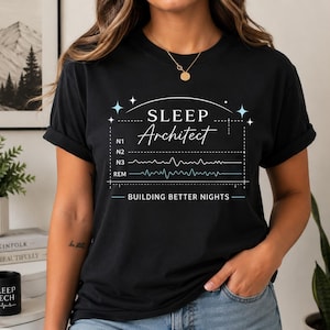 Könnte beinhalten: Ein schwarzes T-Shirt mit weißem und hellblauem Text, der SLEEP Architect lautet, einer Grafik von Schlafphasen-Wellenformen mit der Beschriftung N1, N2, N3 und REM sowie dem Text BUILDING BETTER NIGHTS unter dem Grafikdesign.