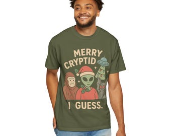Op de afbeelding: Olijfgroen t-shirt met de tekst "MERRY CRYPTID I GUESS." De afbeelding toont een Bigfoot, een alien met een kerstmuts, een UFO en een grijs wezen. Het shirt heeft een ronde hals.