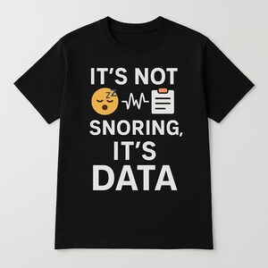 以下が含まれることがあります： 白い文字で「いびきではなく、データです」と書かれた黒いTシャツ。テキストの上に、睡眠のシンボルと折れ線グラフが描かれた黄色の漫画の顔があります。グラフの右側に白いクリップボードのアイコンがあります。