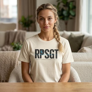Könnte beinhalten: Ein cremefarbenes T-Shirt mit dem schwarzen Text "RPSGT", einem blauen Wellenform- und Monddesign. Darunter steht der Text "POLYSOMNOGRAPHIC TECH". Das Shirt besteht aus einem weichen Material.