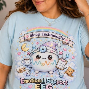 Könnte beinhalten: Ein hellblaues T-Shirt mit einer Grafik einer Zeichentrickfigur, die eine Schlafmütze und einen Stirnspiegel trägt und eine Kaffeetasse sowie ein Klemmbrett hält. Der Text lautet Sleep Technologist, Emotional Support EEG und Always Here For You.