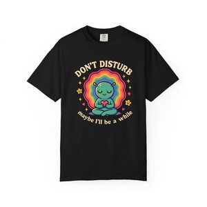 Könnte beinhalten: Schwarzes T-Shirt mit einem farbenfrohen Aufdruck. Der Aufdruck zeigt einen meditierenden Alien, der ein Herz hält, umgeben von einem Regenbogen und Sternen. Der Text lautet "DON'T DISTURB maybe I'll be a while."