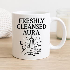 May include: White ceramic mug with the text "FRESHLY CLEANSED AURA" in black. The mug features a black illustration of a hand holding a crystal, with stars, a crescent moon, and radiating lines.