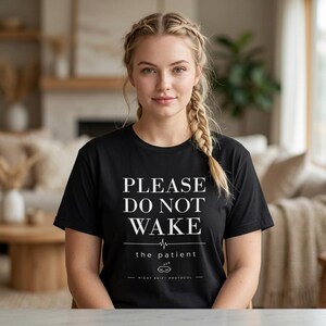 Könnte beinhalten: Schwarzes T-Shirt mit weißem Text "PLEASE DO NOT WAKE the patient" und einer Herzschlag-Grafik. Auf der Unterseite des Shirts steht "NIGHT SHIFT PROTOCOL". Das Shirt wird von einer Person mit blonden Haaren getragen.