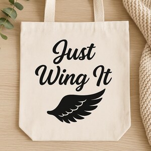 May include: Cream-colored tote bag with the phrase "Just Wing It" in black script above a black wing graphic. The bag has matching cream-colored handles and is made of a sturdy fabric.