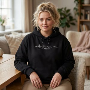 May include: Black hoodie with a drawstring hood and a front pocket. The hoodie features a white graphic of a heartbeat line with the text "Your Name Here" and "RPSGT" below it. The model is wearing the hoodie.