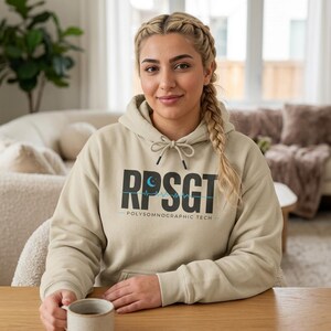 Peut inclure: Un sweat à capuche beige avec le texte "RPSG T" en noir, un croissant de lune et une ligne de battement de cœur. Le texte "POLYSOMNOGRAPHIC TECH" est en dessous. Le sweat à capuche a une capuche à cordon et est porté par une personne.