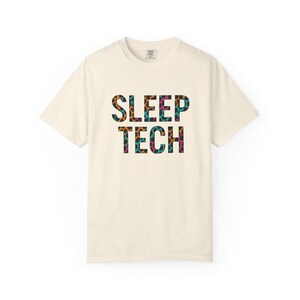 Puede incluir: Una camiseta de color crema con un estampado gráfico de las palabras "SLEEP TECH" en un patrón de estampado de leopardo con acentos naranja, verde azulado y rosa.