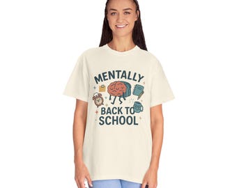 May include: Cream-colored t-shirt with the text "MENTALLY BACK TO SCHOOL" and a graphic of a brain, backpack, alarm clock, notebook, and mug. The design is in shades of brown, blue, and green. The shirt is a comfortable, casual style.