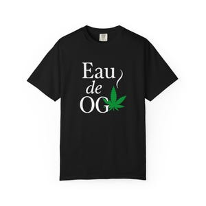 May include: Black t-shirt with the text "Eau de OG" in white, featuring a green cannabis leaf graphic. The shirt is a classic crew neck style.