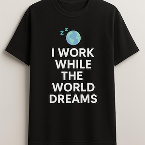 May include: Black t-shirt with a white graphic that says "I work while the world dreams". The graphic also includes a blue and green globe with zzz's above it.