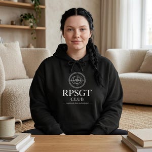 Peut inclure: Un sweat à capuche noir avec le texte blanc "RPSGT CLUB" et un graphisme circulaire. Le texte "registered sleep technologist" est en dessous du nom du club. La personne est assise à une table en bois.