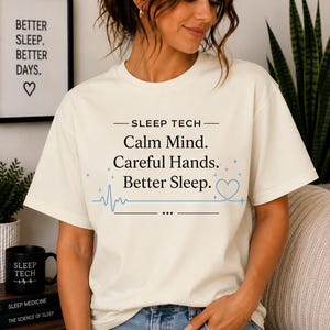 Peut inclure: Un t-shirt à manches courtes de couleur crème avec le texte noir "SLEEP TECH" suivi de "Calm Mind. Careful Hands. Better Sleep." et un motif bleu clair représentant un cœur et une ligne de pouls avec de petites étoiles sur la poitrine.