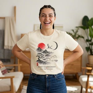 May include: A cream-colored short-sleeve t-shirt featuring a black and red ink-style graphic of mountains, a bonsai tree, a crane, and a crescent moon. The text reads SLEEP TECHNOLOGIST and Quieter Minds, Better Nights, with a small red stamp.