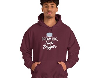 Könnte beinhalten: Ein weinrotes Kapuzensweatshirt mit einer Fronttasche. Das Sweatshirt zeigt den Text "DREAM BIG, Nap Bigger" in Weiß und eine hellblaue Kissen-Grafik. Das Model trägt eine blaue Jeans.