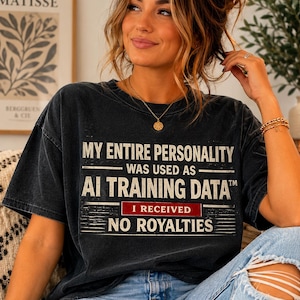 May include: A black, vintage-wash short-sleeve t-shirt featuring white text that reads: MY ENTIRE PERSONALITY WAS USED AS AI TRAINING DATA I RECEIVED NO ROYALTIES. The text includes a red rectangular accent box around the words I RECEIVED.
