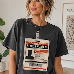 May include: A charcoal gray short-sleeve t-shirt featuring a graphic of a beige identification badge. The badge text reads Human Resources, Senior Human, Level 37, Organic Intelligence, Firmware: Childhood Trauma, Battery: Coffee, Status: Still Operational, and ID# 042786.